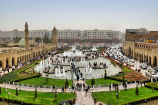 Erbil / Hewler / Arbil / Irbil, Kurdistan, Iraq: main square, Shar Park, with crowds enjoying the pleasantly cool area created by the fountains - arcades on both sides and Nishtiman mall in front - Mosque and Erbil Clocktower on the left - dense traffic on Kirkuk avenue on the right - seen from the Erbil citadel - photo by M.Torres