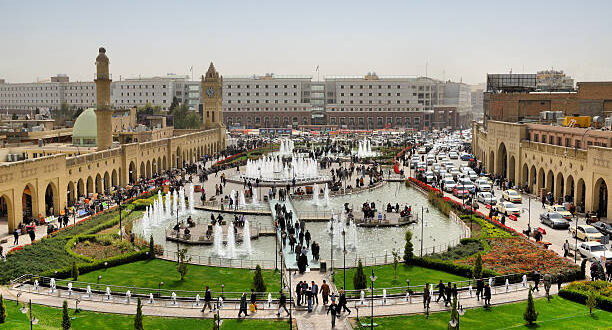 Erbil / Hewler / Arbil / Irbil, Kurdistan, Iraq: main square, Shar Park, with crowds enjoying the pleasantly cool area created by the fountains - arcades on both sides and Nishtiman mall in front - Mosque and Erbil Clocktower on the left - dense traffic on Kirkuk avenue on the right - seen from the Erbil citadel - photo by M.Torres